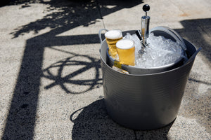 Two glasses of beer in a large metal tub filled with ice on a concrete surface.